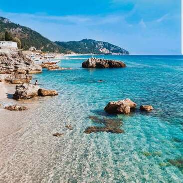 The image depicts a serene beach scene with crystal-clear water, rocky shoreline, and distant mountains under a blue sky with scattered clouds.