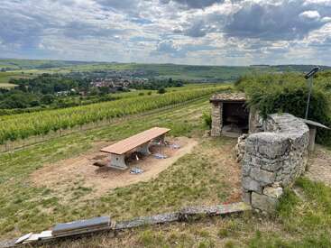 Un banc de pique-nique en bois trône au milieu de vignobles verdoyants, avec vue sur un village lointain. Un ancien abri en pierre et un mur ajoutent une touche rustique sous un ciel nuageux spectaculaire.