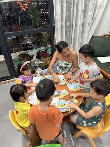 A woman sits at a small round table with five children, helping them with colorful activity books. The atmosphere appears lively and focused in a cozy room.