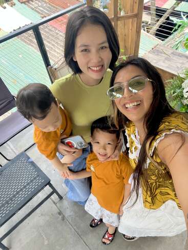 Two women and two young children pose happily for a selfie outdoors. Everyone is smiling, wearing casual clothing, and the children wear matching orange shirts. Warm, cheerful atmosphere.