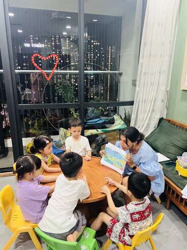 A woman reads a colorful storybook to five attentive children seated around a small table. City lights and a heart shape glow outside the window.