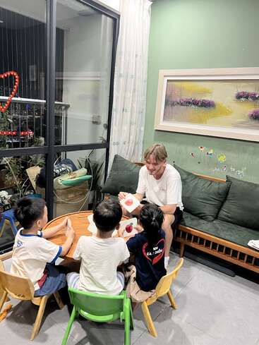 Un hombre se sienta con tres niños pequeños en una pequeña mesa, enseñándoles con fichas. La acogedora habitación está decorada con plantas, arte y pegatinas.