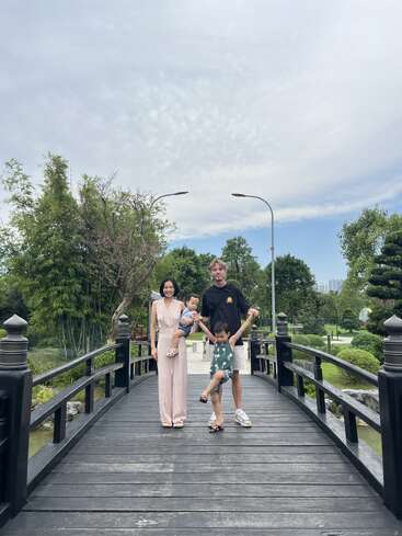 Una familia de cuatro miembros posa sobre un puente de madera en un frondoso y verde parque bajo un cielo parcialmente nublado, sonriendo y disfrutando juntos de un tranquilo momento al aire libre.