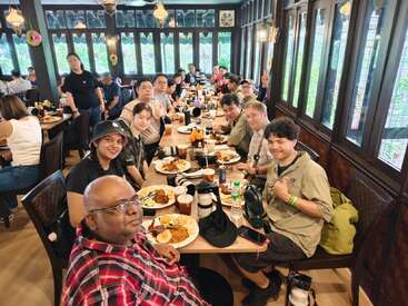 Un grand groupe de personnes est assis autour d'une longue table dans un restaurant ; elles dégustent un repas et des boissons, sourient et discutent dans un cadre chaleureux et bien éclairé.