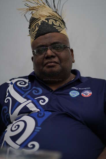 Un homme à lunettes porte un chapeau traditionnel tissé orné de plumes et une chemise à motifs bleus. Assis devant un fond uni, il semble calme et digne.