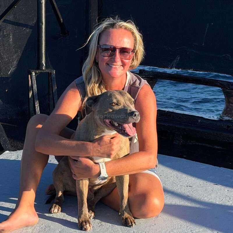 Eine lächelnde Frau sitzt barfuß im Freien und umarmt einen fröhlichen braunen Hund. Beide wirken entspannt und zufrieden. Im Hintergrund sind dunkle Gebäude und ein Stück Wasser zu sehen.