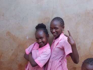 Zwei lächelnde Mädchen stehen an einer Wand und posieren spielerisch. Beide tragen rosa karierte Uniformen. Die eine verschränkt ihre Arme, während die andere ein Friedenszeichen macht. Ein fröhlicher Moment.