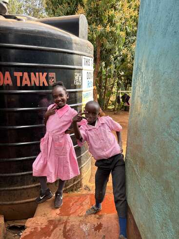 Zwei lächelnde Kinder in rosa Schuluniformen posieren spielerisch neben einem großen schwarzen Wassertank und einer türkisfarbenen Wand, draußen an einem sonnigen Tag mit Bäumen in der Nähe.
