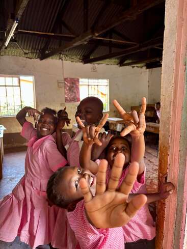 Vier fröhliche Kinder in rosafarbenen Schuluniformen posieren spielerisch in einem rustikalen Klassenzimmer, lächeln und machen Handgesten und strahlen gemeinsam Glück und Aufregung aus.