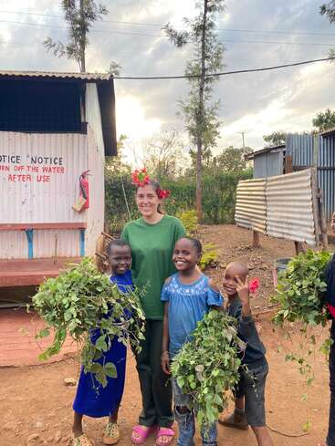 Eine lächelnde Frau mit Blumenschmuck und drei glückliche Kinder, die grüne Pflanzen halten, stehen vor einem Metallgebäude, umgeben von Bäumen unter einem bewölkten Himmel.