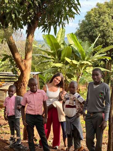 Eine Gruppe von Kindern und eine Frau posieren im Freien zwischen grünen Bäumen und Pflanzen, lächeln und genießen einen sonnigen Tag, tragen Freizeit- und Schulkleidung.
