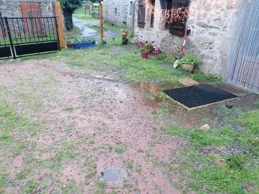 Un patio húmedo y embarrado con charcos, macetas, guirnaldas de luces, un felpudo negro, una casa de piedra, una verja metálica y vegetación dispersa tras la lluvia.