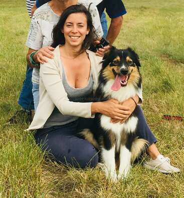Das Bild zeigt eine Frau, die auf einer Wiese sitzt und einen großen Hund umarmt, dessen Arme sie um seine Brust geschlungen hat. Der Hund hechelt und scheint glücklich zu sein.
