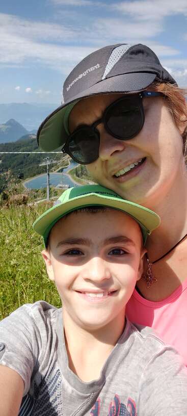 Una mujer y un niño sonrientes posan para hacerse un selfi al aire libre, con gorras y gafas de sol. Detrás de ellos se ve un pintoresco paisaje montañoso con un lago y un cielo azul.