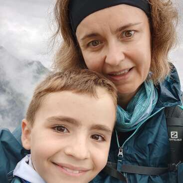 Una mujer y un niño pequeño sonríen muy juntos para hacerse un selfi. Llevan chaquetas de montaña, y al fondo se ven montañas envueltas en niebla y un cielo nublado.