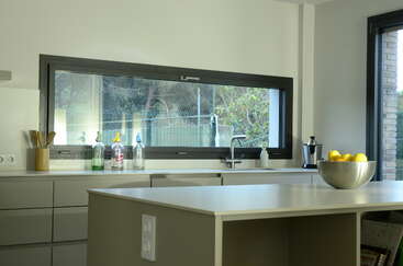 Esta cocina moderna cuenta con un gran ventanal horizontal, armarios minimalistas, una encimera elegante, cuencos con limones, botellas de cristal, una cafetera y mucha luz natural.