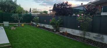 Un jardín trasero muy bien cuidado, con césped artificial, arbustos floridos, árboles y plantas en macetas a lo largo de una valla oscura. Al fondo se ven casas y árboles.