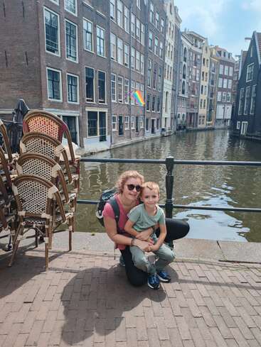 Una mujer y un niño sonríen junto a un canal en una ciudad europea. En el fondo se ven sillas apiladas, una bandera arcoíris y edificios altos con muchas ventanas.