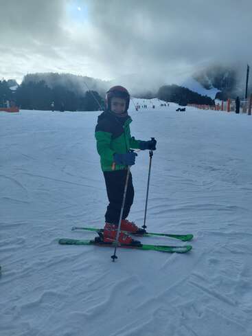 Un niño con chaqueta verde y casco rojo está de pie sobre los esquís, sonriendo en una pista nevada. Al fondo se ven montañas envueltas en niebla, árboles y esquiadores en la lejanía.