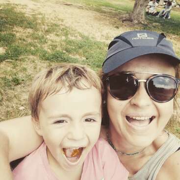 Una niña alegre y una mujer adulta sonríen ampliamente mientras se hacen un selfi al aire libre. La mujer lleva gafas de sol y una gorra, con césped verde y árboles al fondo.