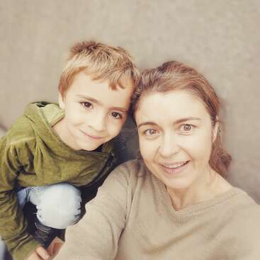 A smiling woman and young boy pose closely for a selfie. Both look happy, with the boy wearing a green hoodie and the woman in a beige sweater.