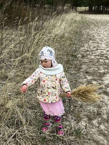 Ein kleines Kind in bunter, gemusterter Kleidung steht auf einem Feld, hält einen Strauß getrocknetes Gras in der Hand und ist von Natur und hohem, trockenem Schilf umgeben.