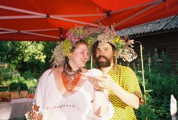 Ein fröhliches Paar mit bunten Blumenkränzen und festlicher Kleidung steht unter einem roten Baldachin und genießt bei sonnigem Wetter gemeinsam im Freien ein Stück Kuchen.