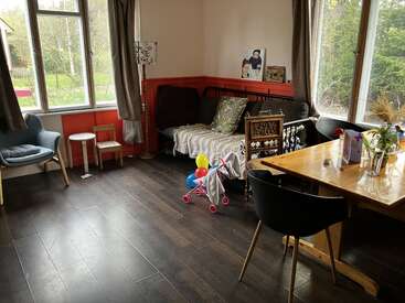 Ce salon chaleureux est doté de grandes fenêtres, d'un parquet, d'un canapé, de jouets pour enfants, d'une table à manger avec des chaises, de plantes et bénéficie d'une belle luminosité naturelle.