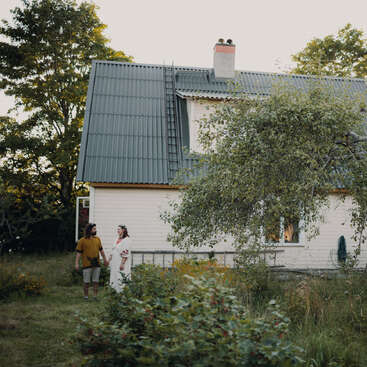 Un couple se tient par la main dans un jardin verdoyant et luxuriant, devant une maison blanche chaleureuse au toit métallique, entourée d'arbres et de plantes.