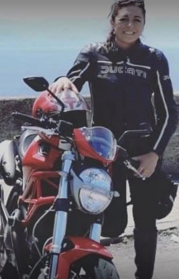 A woman wearing a black Ducati jacket stands next to a red motorcycle. She smiles at the camera, holding a helmet, with a blue sky background.