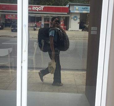 Uma pessoa com uma mochila grande e uma sacola plástica tira uma selfie no espelho de uma porta de vidro. Dá pra ver uma loja de conveniência e a rua ao fundo.
