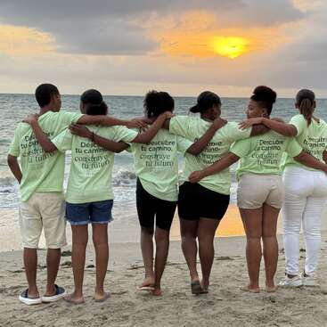 Sechs Menschen stehen Arm in Arm an einem Strand, tragen zusammenpassende T-Shirts mit der Aufschrift "Descubre tu camino, construye tu futuro" und beobachten einen farbenfrohen Sonnenuntergang über dem Ozean.