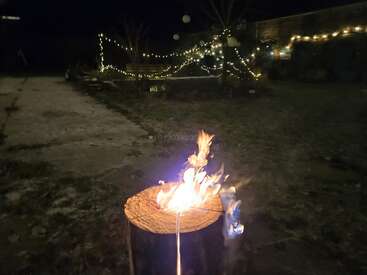Eine gemütliche Nachtszene im Freien mit einem brennenden Feuer in einem Baumstamm, festlich drapierten Lichterketten im Hintergrund und einer friedlichen, leicht frostigen Gartenlandschaft.