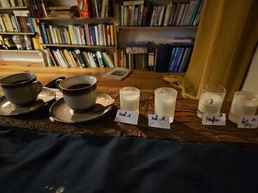 Zwei Kaffeetassen auf Untertassen stehen neben vier beschrifteten Kerzen auf einem Holztisch. Bücherregale und ein gelber Vorhang bilden den gemütlichen, warm beleuchteten Hintergrund.