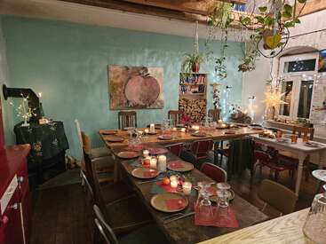 In einem gemütlichen Esszimmer steht ein langer Holztisch, der für das Abendessen gedeckt ist, umgeben von ungleichfarbigen Stühlen, Kerzen, festlicher Dekoration, hängenden Pflanzen und warmer Beleuchtung.