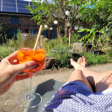 Eine Person entspannt sich an einem sonnigen Tag im Freien und hält einen Cocktail mit Eis und Orangenscheiben in der Hand, umgeben von üppigem Grün und festlichen hängenden Laternen.