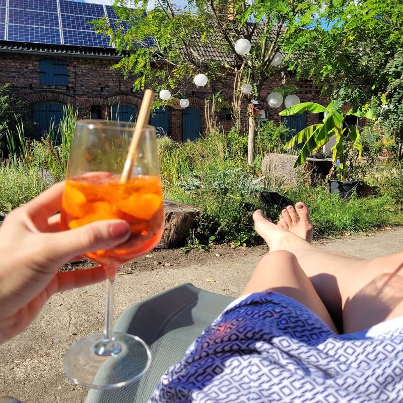 Eine Person entspannt sich an einem sonnigen Tag im Freien und hält einen Cocktail mit Eis und Orangenscheiben in der Hand, umgeben von üppigem Grün und festlichen hängenden Laternen.