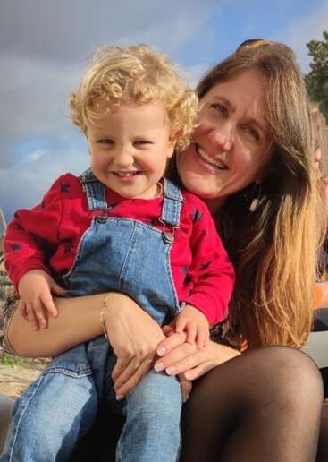 The image depicts a woman with long brown hair and a child with blonde curly hair, posing together in an outdoor setting, likely a park or garden.
