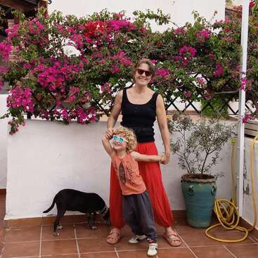 Eine lächelnde Frau und ein Kind mit Sonnenbrillen posieren fröhlich auf einer Terrasse mit rosa Blumen, einer Topfpflanze, einem gelben Schlauch und einem kleinen schwarzen Hund.
