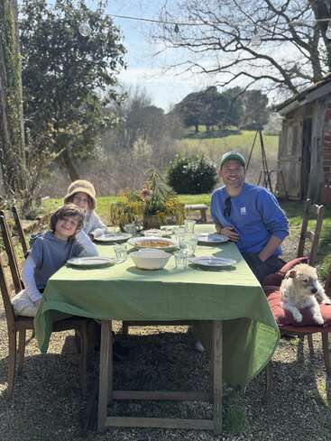A family enjoys an outdoor meal on a sunny day. Three people and a dog sit around a table with food, surrounded by nature and trees.