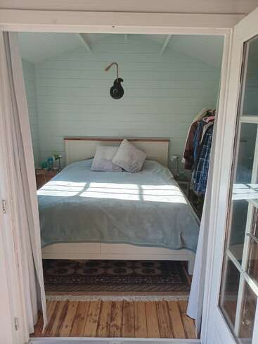 A cozy, sunlit bedroom with a double bed, light-colored walls, two pillows, wooden floors, a patterned rug, clothes rack, and decorative wall hanging above the bed.