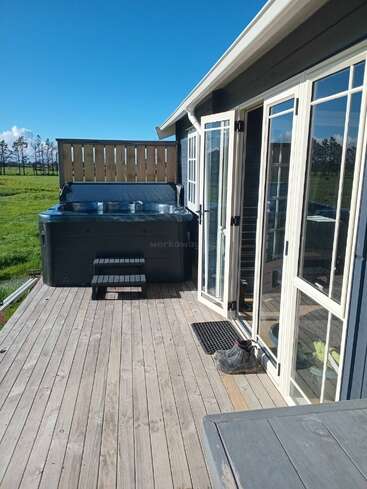 A wooden deck with a hot tub, boots, and open glass doors, overlooks a green field under a clear blue sky, creating a relaxing outdoor scene.