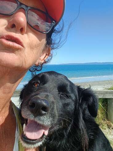 Une femme portant des lunettes de soleil et une casquette orange pose avec un chien noir joyeux près de la plage. Le ciel est dégagé et l'océan est magnifique.
