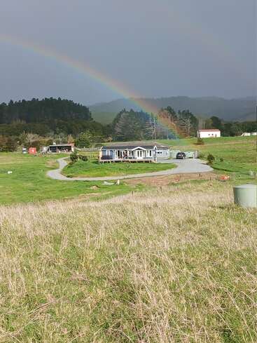 Une maison pittoresque se trouve dans des champs verdoyants sous un ciel nuageux, avec un double arc-en-ciel éclatant au-dessus, entourée d'arbres, de collines et de bâtiments ruraux.
