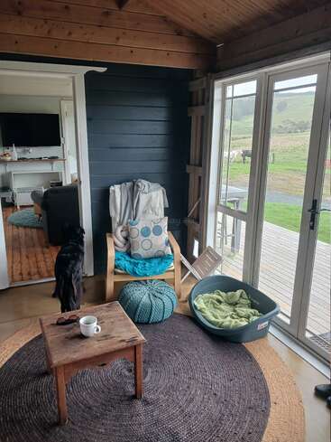 Une véranda confortable avec un plafond en bois, un fauteuil confortable, un pouf et un panier pour chien. Un chien noir se tient près de la porte. De grandes fenêtres offrent une vue panoramique sur la campagne.