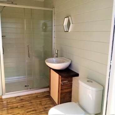 Das ist ein modernes Badezimmer mit einer Glasdusche, Holzboden, einem Waschbecken auf einem Holzunterschrank, einer Toilette, hellen Wandpaneelen und einem sechseckigen Spiegel.