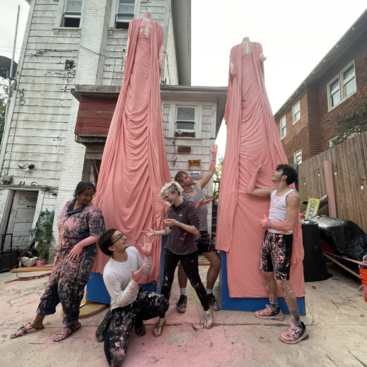 A imagem mostra um grupo de cinco pessoas posando em frente a duas esculturas altas de tecido rosa em um ambiente urbano, cercado por prédios e detritos.