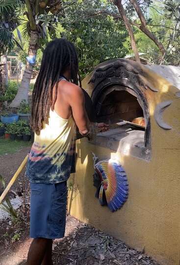 A person with long dreadlocks bakes bread in a rustic outdoor oven decorated with colorful Native-inspired art, surrounded by lush green trees and plants.