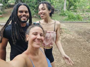 Three smiling people pose for a cheerful selfie outdoors, surrounded by lush green vegetation, trees, and a garden. They appear relaxed, happy, and enjoying time together.