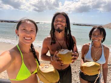 Three friends smile on a sunny beach, holding fresh coconuts. The ocean and blue sky with clouds form a beautiful, relaxing tropical background. Summer vibes abound.
